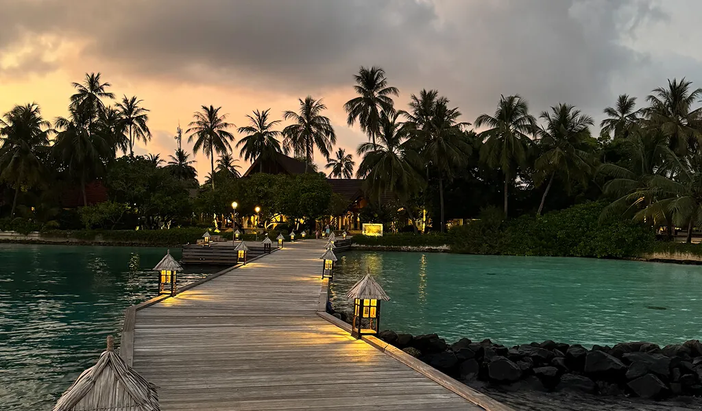 Holzsteg mit Laternen führt ins Resort auf den Malediven, umgeben von Palmen und türkisfarbenem Wasser